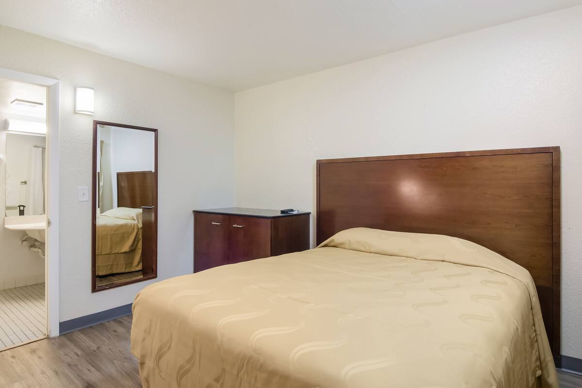 A hotel room with a wooden headboard, a bed with a beige patterned bedspread, a small wooden dresser, and a mirror on the wall.