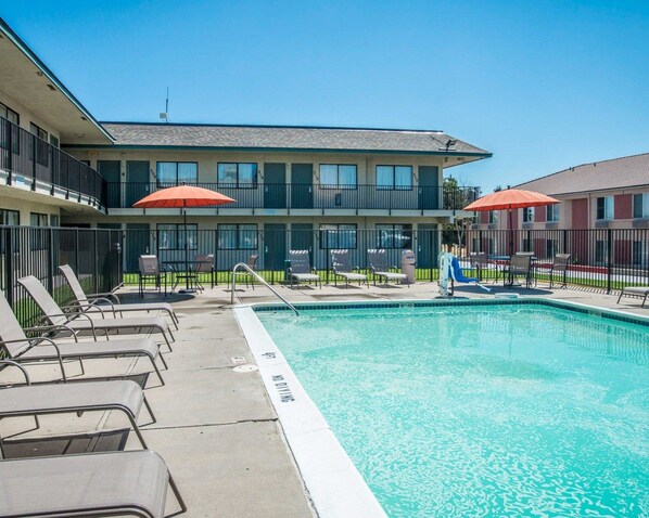 A swimming pool with lounge chairs and umbrellas, surrounded by a fenced area with a building in the background.