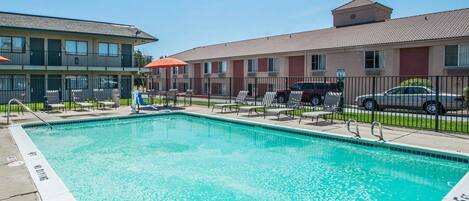A swimming pool with lounge chairs and umbrellas, surrounded by a fence and buildings.
