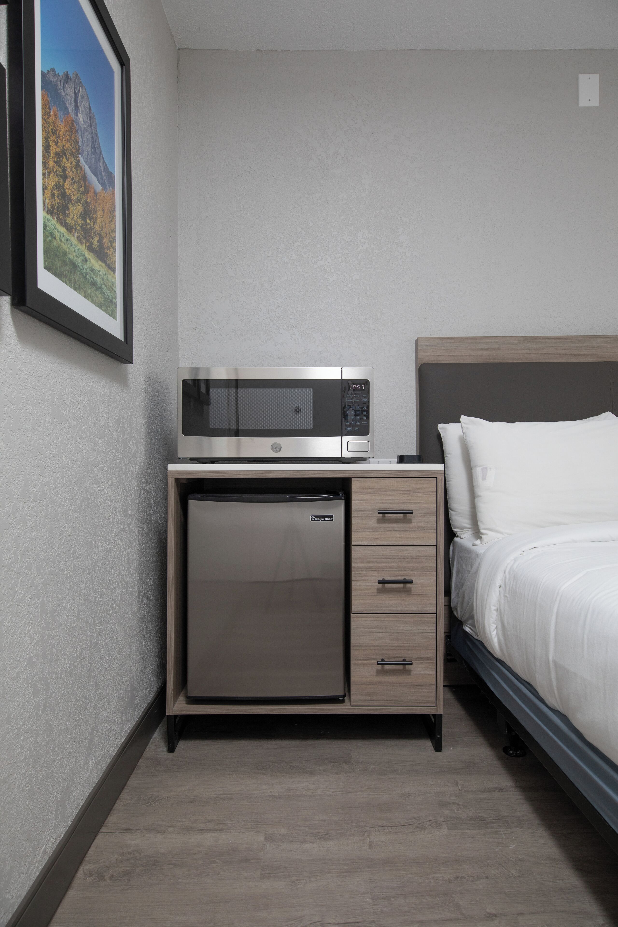 Camera d'albergo compatta con un letto, un minibar, una piccola scrivania con un forno a microonde e un quadro appeso al muro.