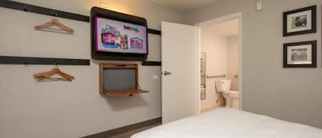 Habitación de hotel con una cama, un televisor montado en la pared, un estante de madera y una puerta que da al baño.