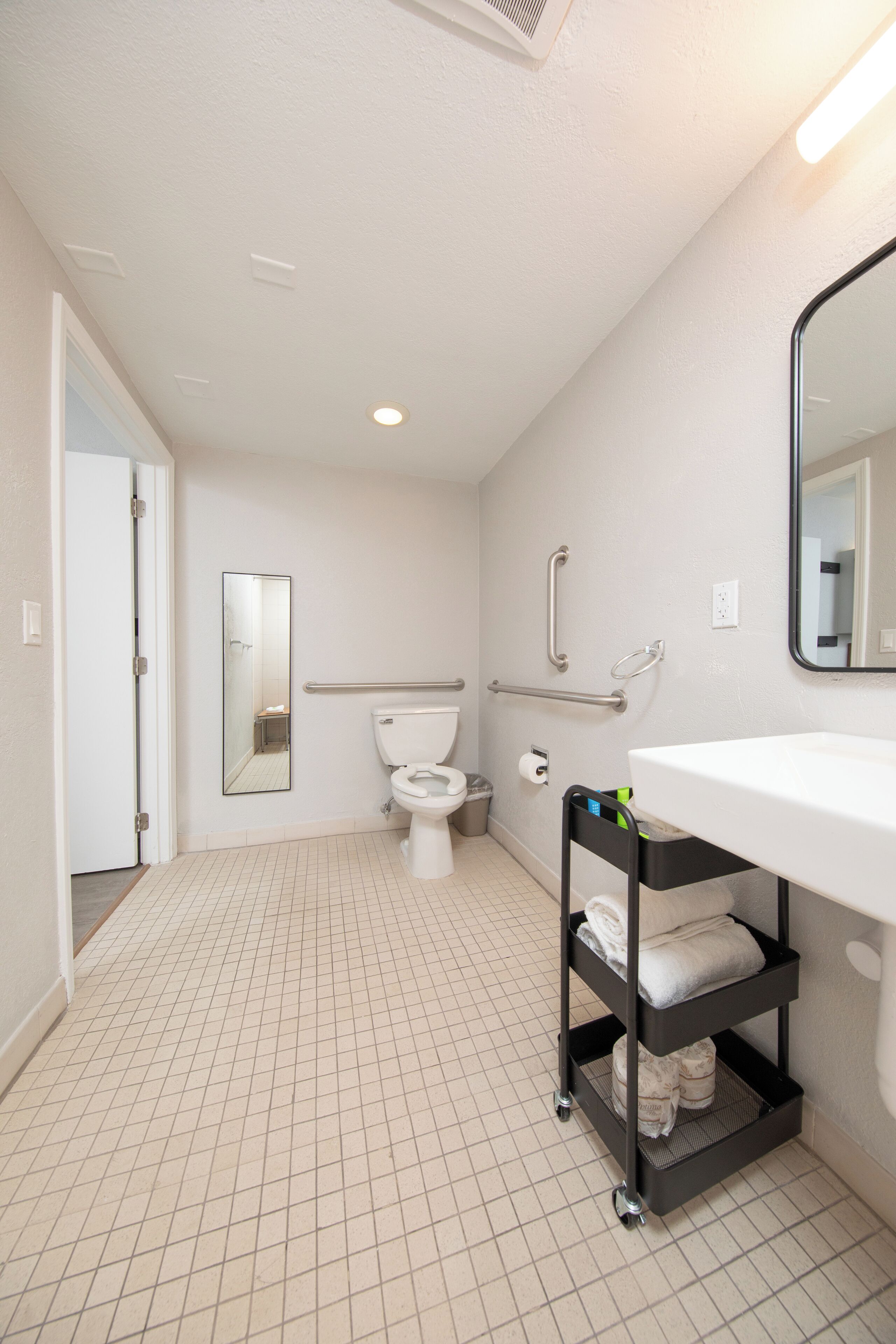 A bathroom with a toilet, a sink, a mirror, a handrail, and a shelf with towels.