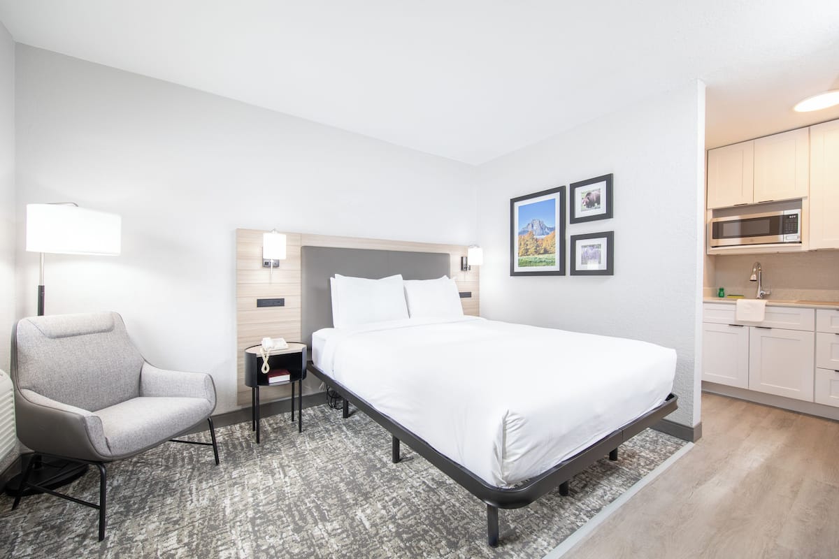 A modern hotel room with a bed, a chair, a nightstand, a lamp, and a kitchenette.