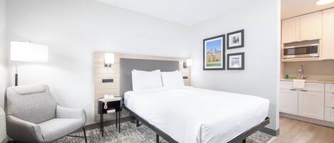 A modern hotel room with a bed, a chair, a nightstand, a lamp, and a kitchenette.
