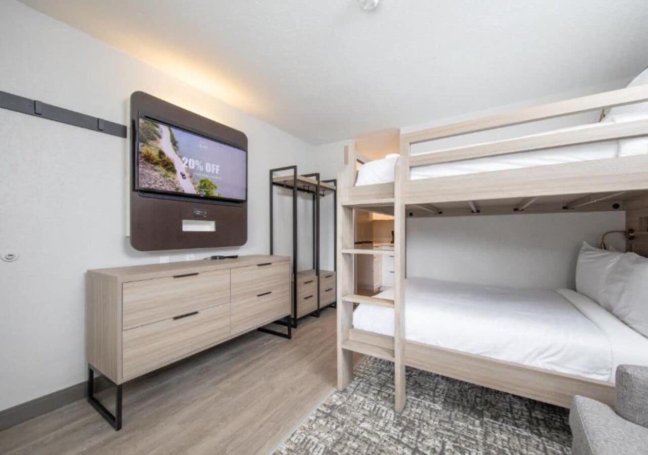 A modern hotel room with a bunk bed, a TV mounted on the wall, a wooden dresser, and a patterned rug.