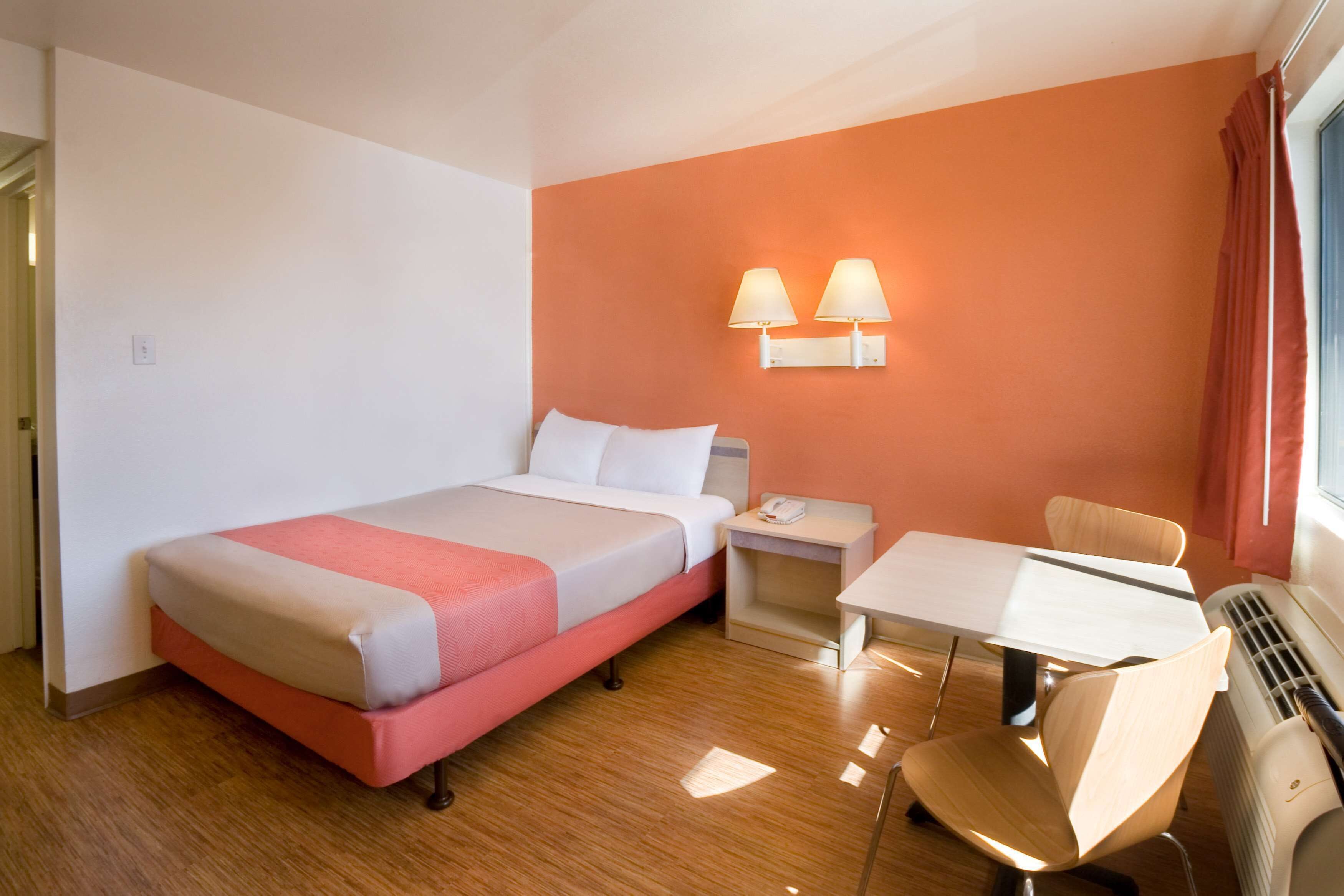 A hotel room with a bed, bedside table, chair, table, and a wall-mounted lamp.