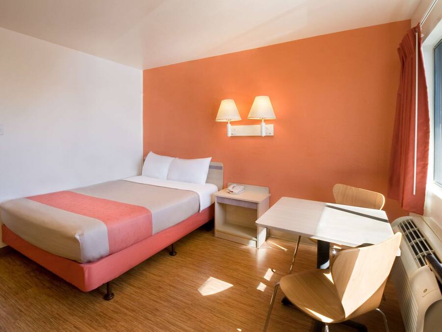 A hotel room with a bed, bedside table, chair, table, and a wall-mounted lamp.