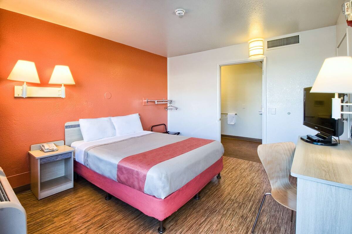 A hotel room with a bed, bedside table, lamp, TV, and a desk with a chair.