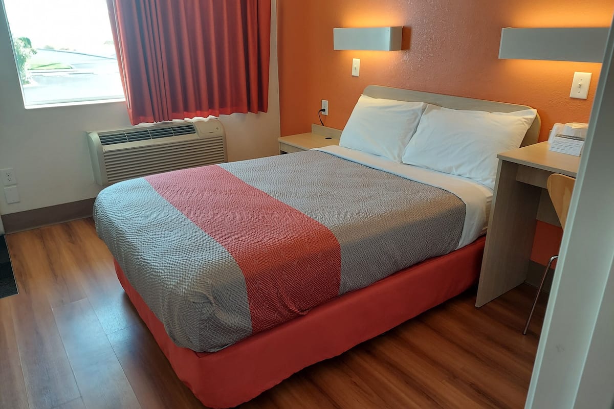 A hotel room with a bed, a desk, a chair, a window with curtains, an air conditioning unit, and a wall-mounted light fixture.