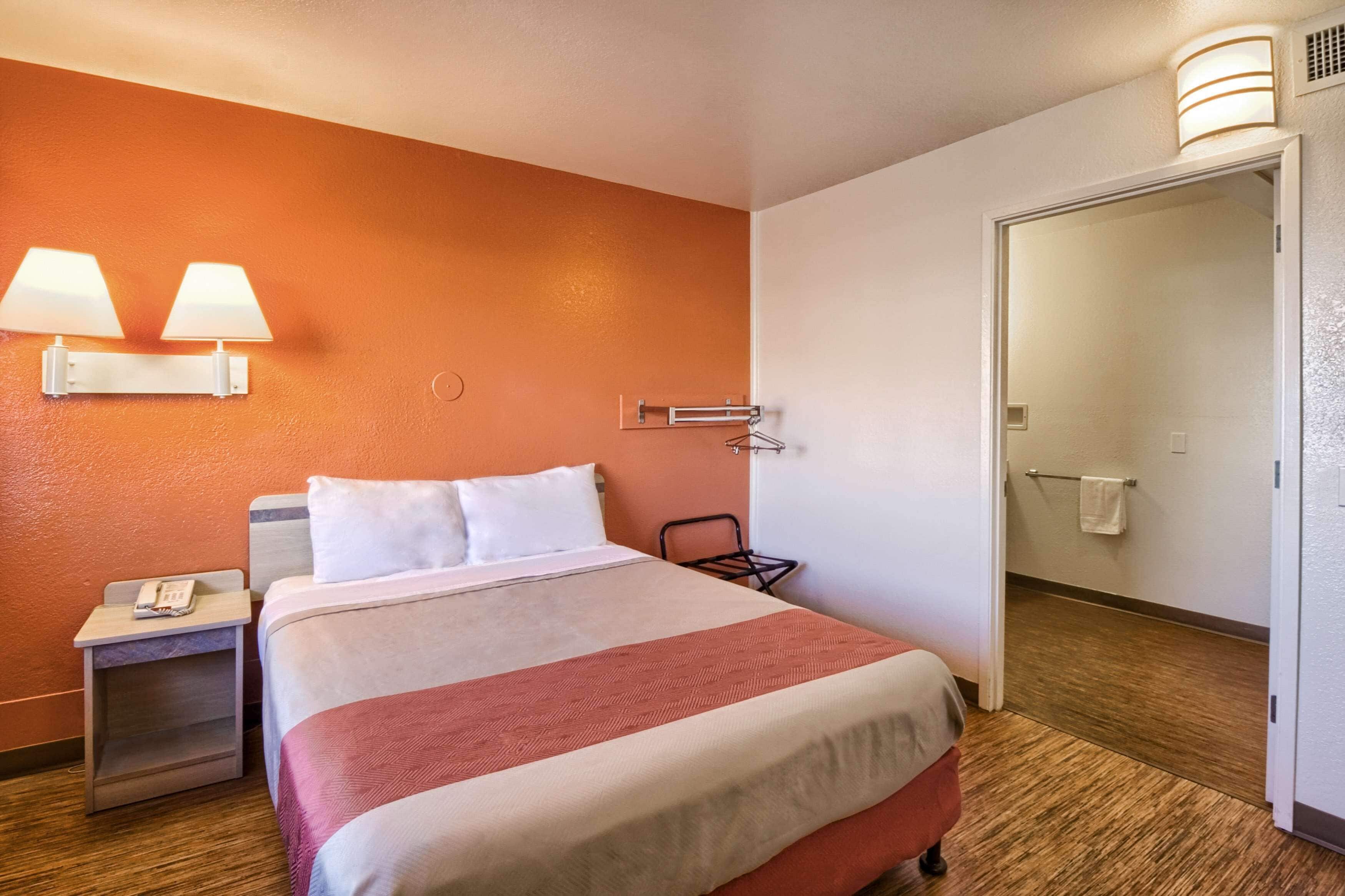 A hotel room with a bed, two bedside lamps, a nightstand, a chair, and a door leading to another room.