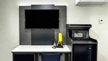 Quarto de hotel compacto com televisão de tela plana na parede, pequeno espaço de cozinha com micro-ondas e mesa com cadeira.
