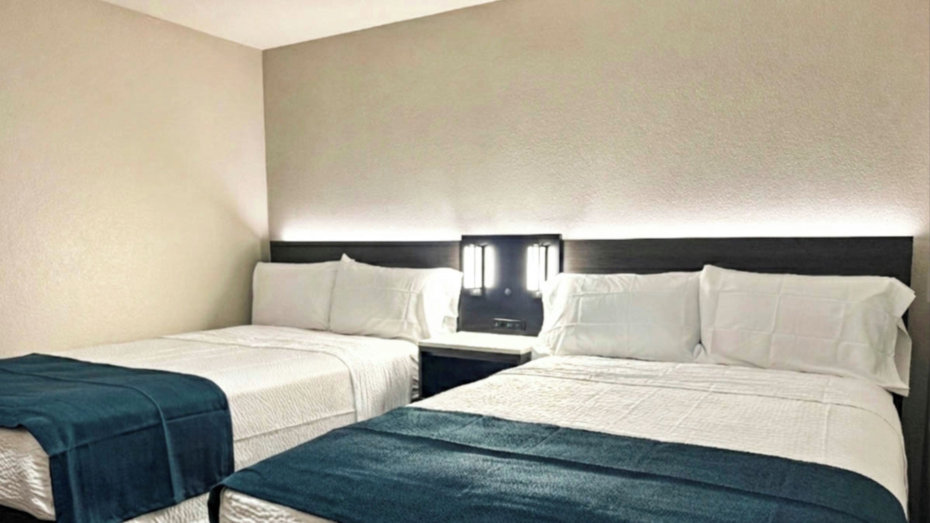A hotel room with two beds, each with white linens and a dark blue blanket. There is a nightstand between the beds with a lamp and a small table.