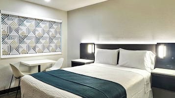 A hotel room with a bed, a desk, and a chair. There is a geometric patterned wall hanging above the desk.