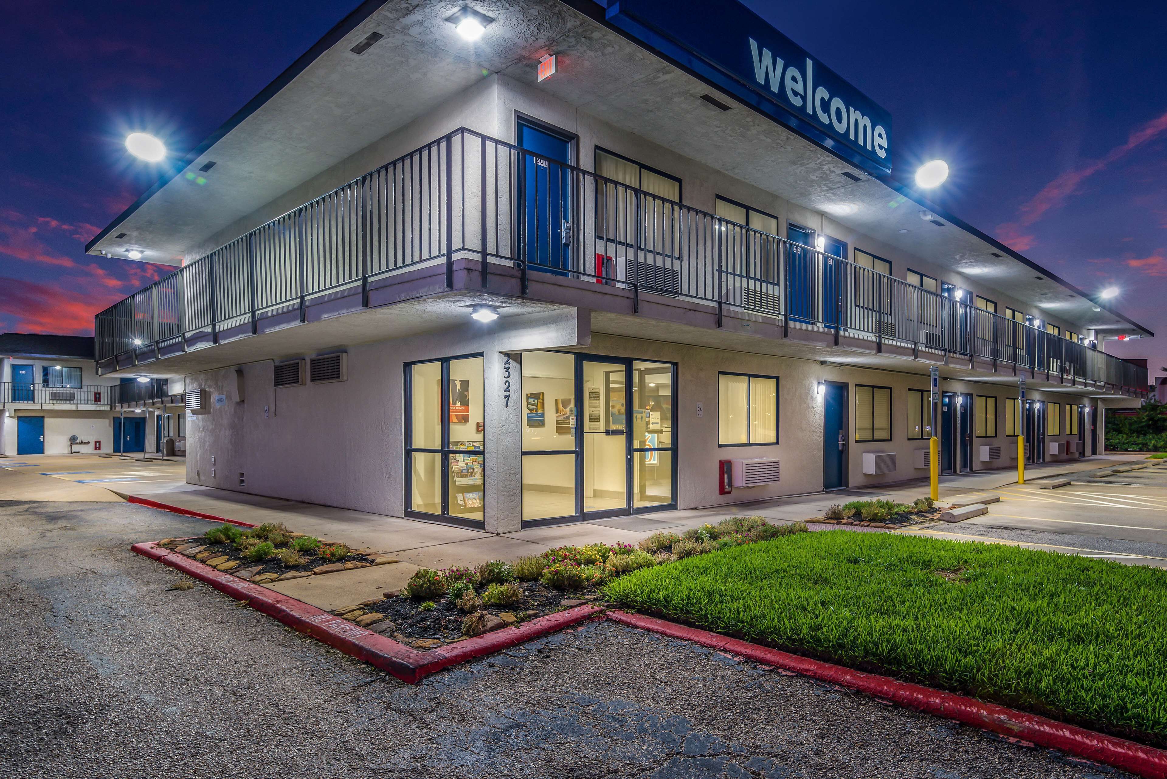 Um motel de dois andares com um letreiro 'Welcome', área de estacionamento coberta e um jardim frontal bem cuidado.