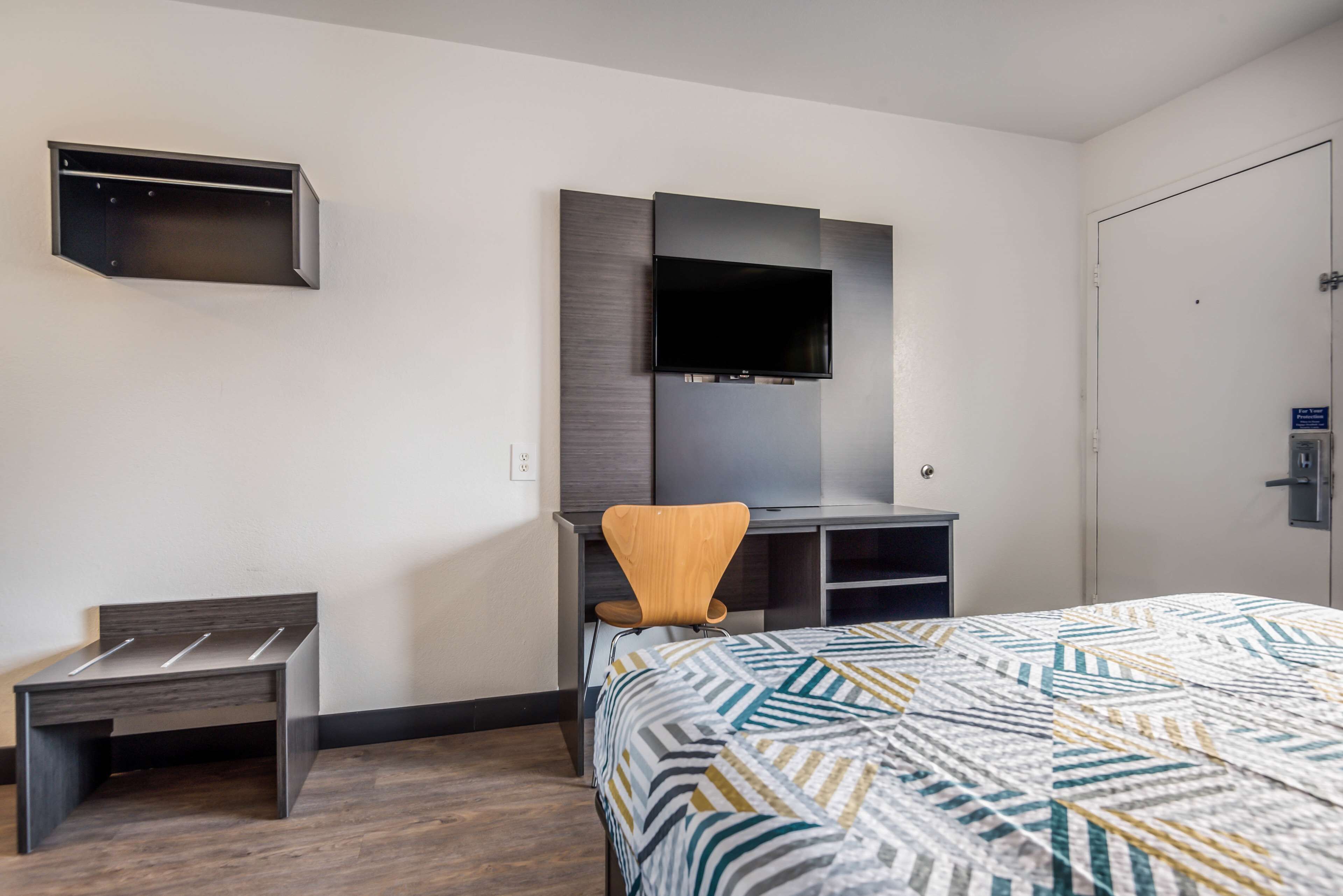 Una habitación de hotel moderna con cama, escritorio, silla, televisor montado en la pared y un pequeño banco.