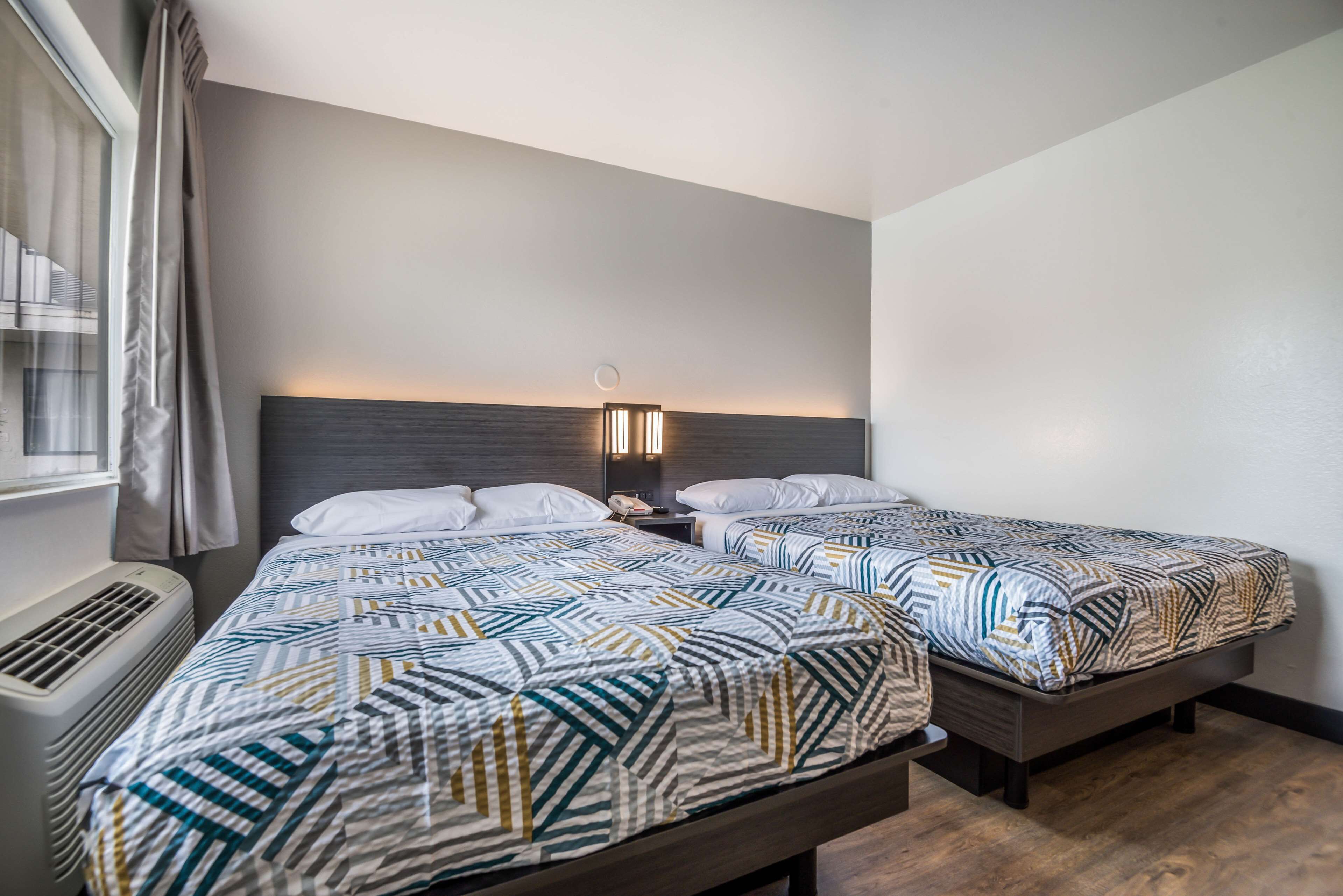 Een hotelkamer met twee bedden, een kachel, een nachtkastje met lamp en een raam met gordijnen.