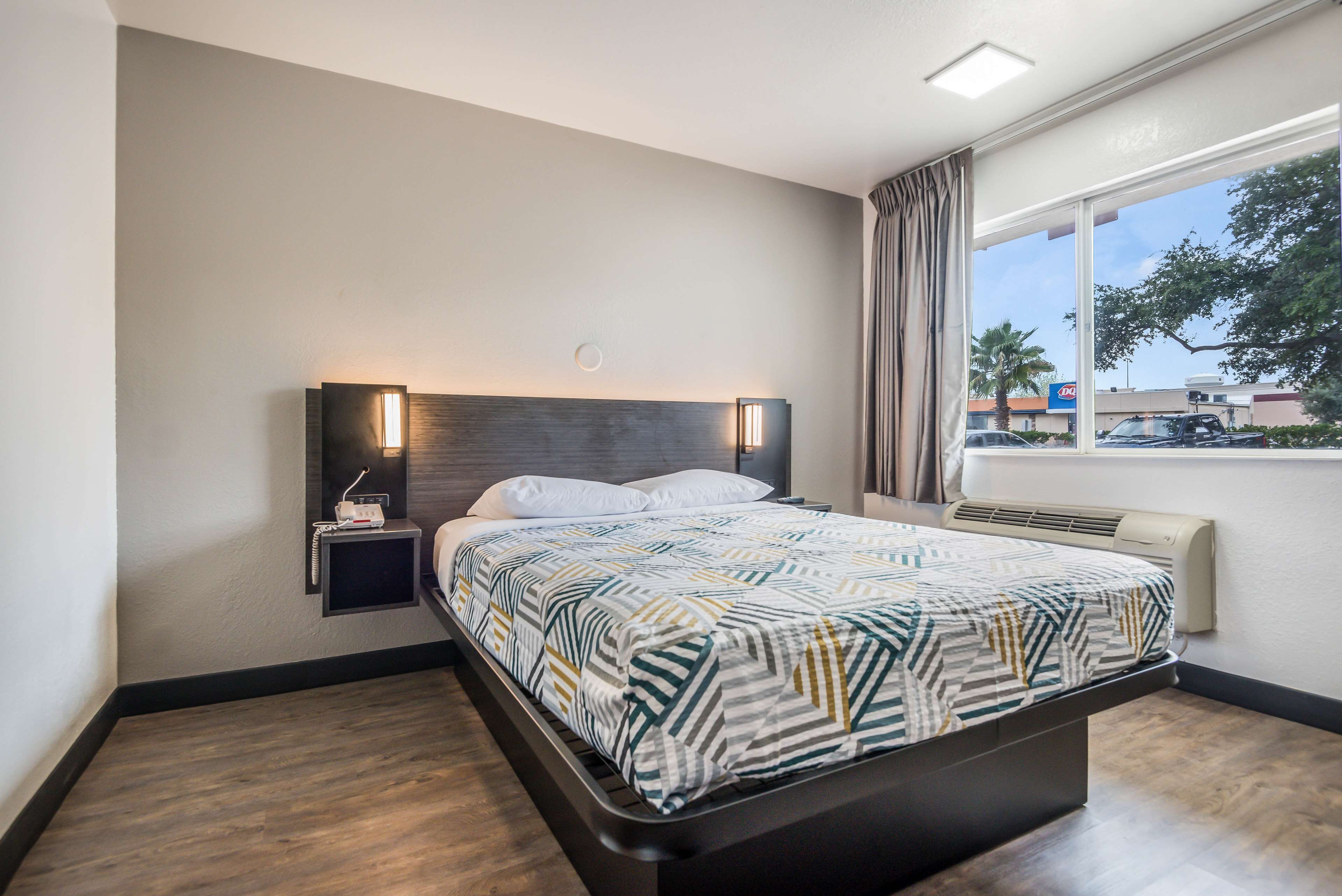 A modern hotel room with a large bed, a bedside table with a lamp, a window with a view of the outside, and a built-in air conditioning unit.
