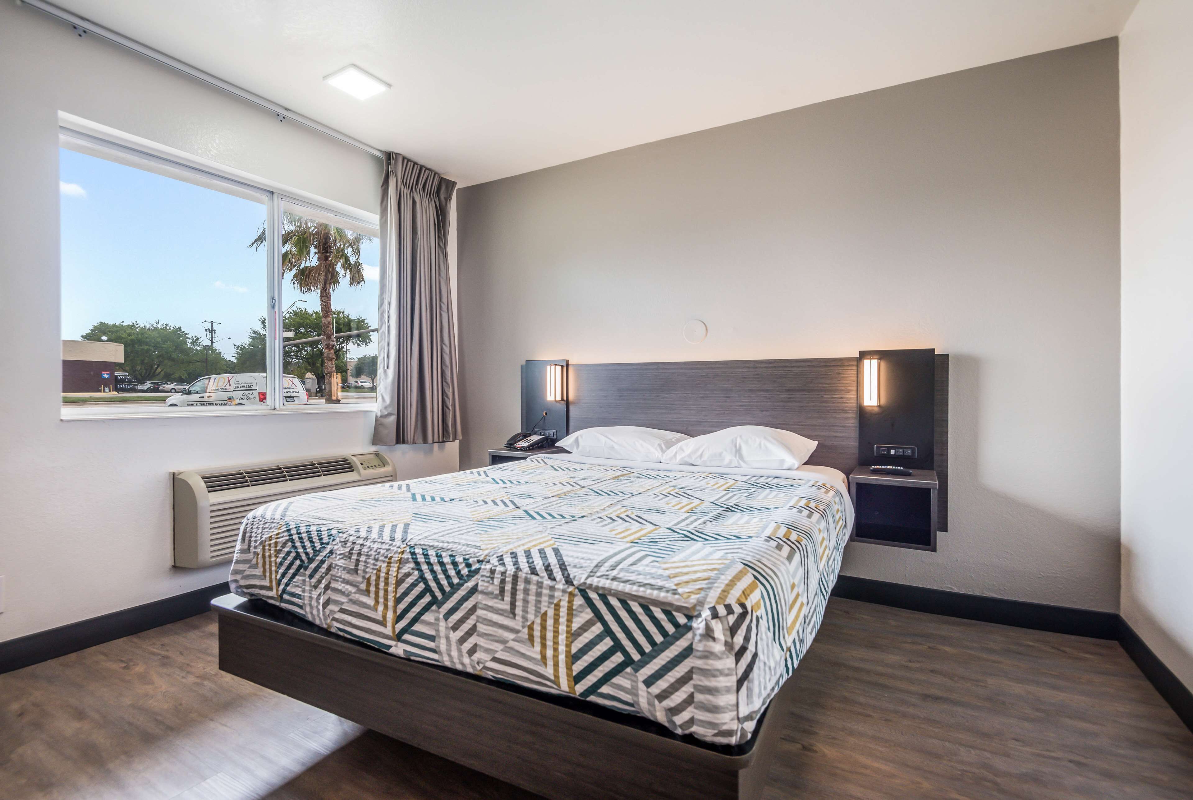 Una habitación de hotel moderna con una cama grande, una mesita de noche con teléfono y lámpara, una ventana con vistas a palmeras y un estacionamiento, y un calentador fijado a la pared.