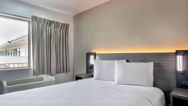 A hotel room with a large bed, a bedside table, a nightstand, a window with curtains, and a wall-mounted light fixture. - Motel 6 Austin, TX - North (Austin)