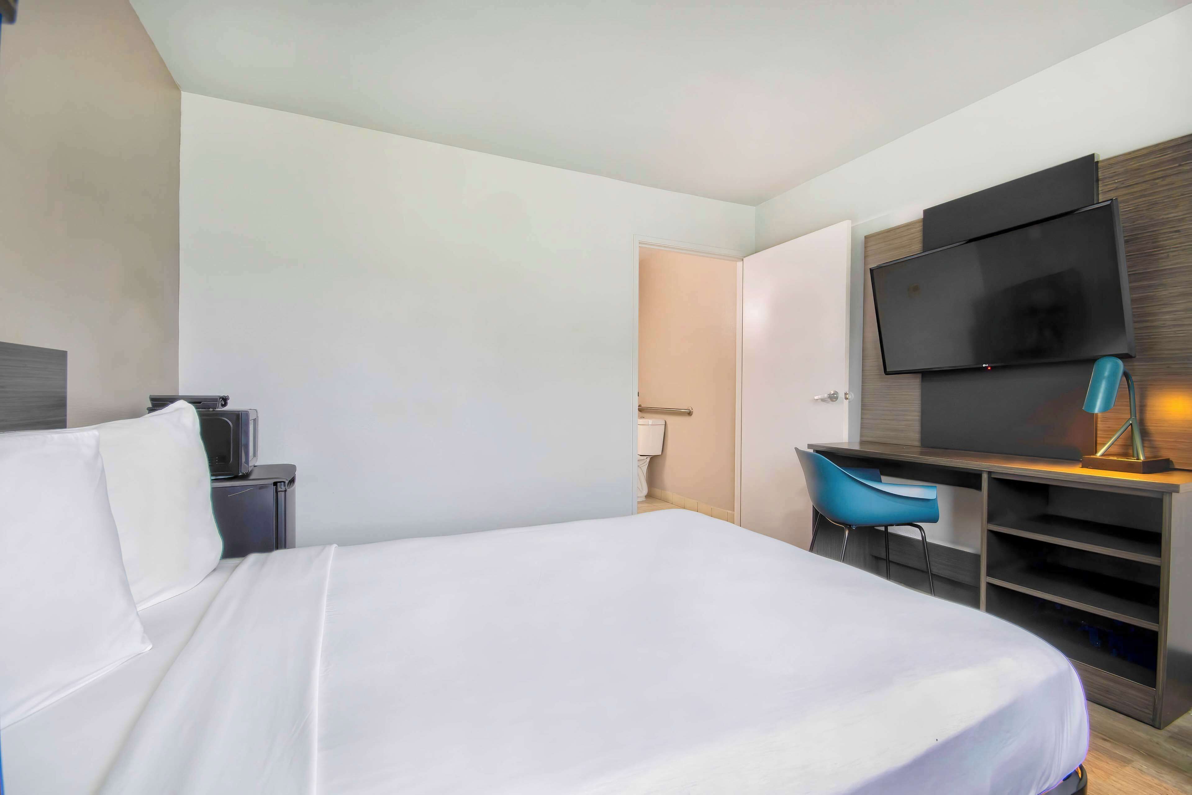 Una habitación de hotel moderna con una cama grande, un escritorio con una silla, un televisor de pantalla plana montado en la pared y una mesita de noche con una lámpara.