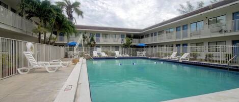 A swimming pool with lounge chairs and umbrellas, surrounded by a multi-story building with balconies.
