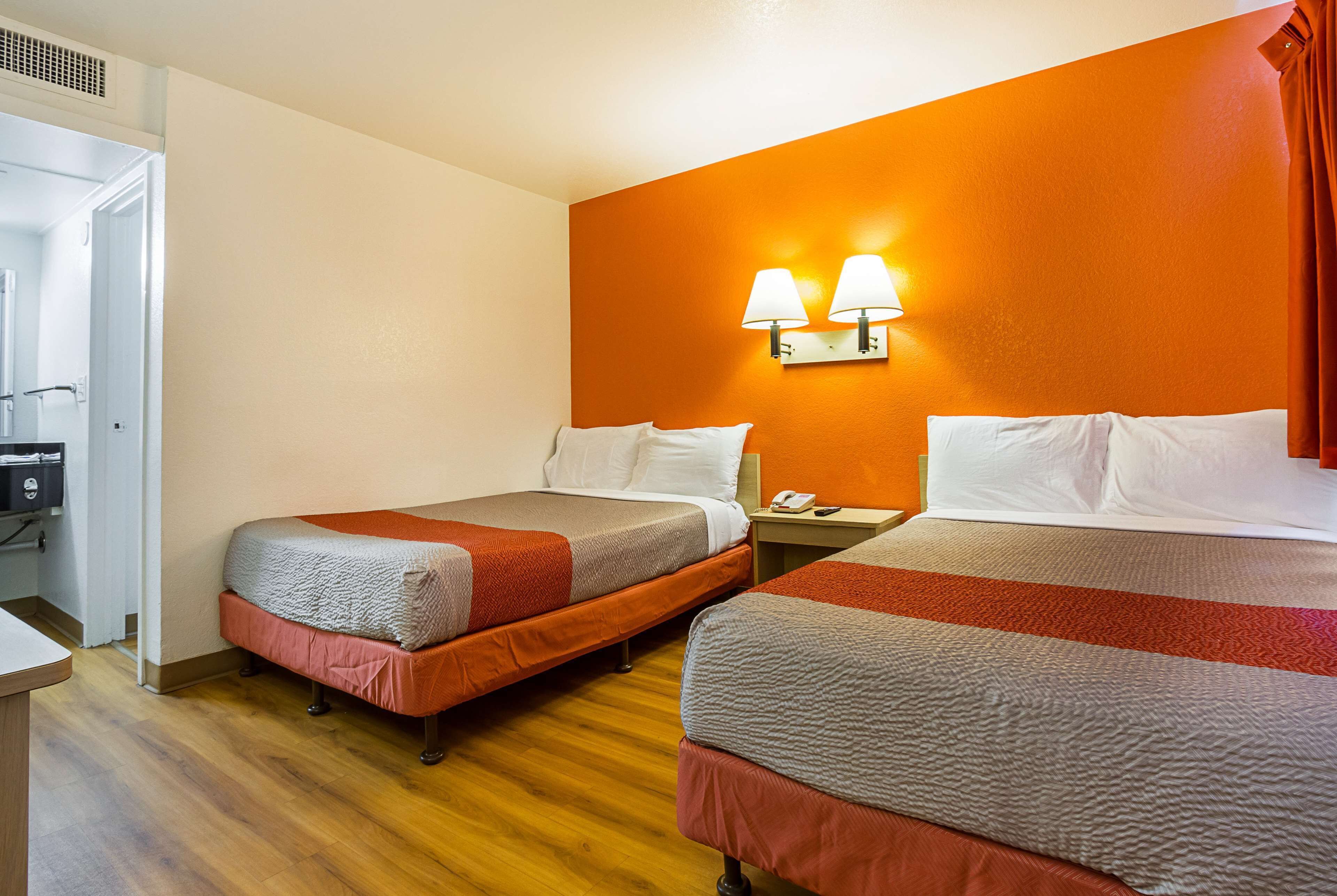 A hotel room with two beds, wooden flooring, an orange accent wall, and a desk with a telephone.