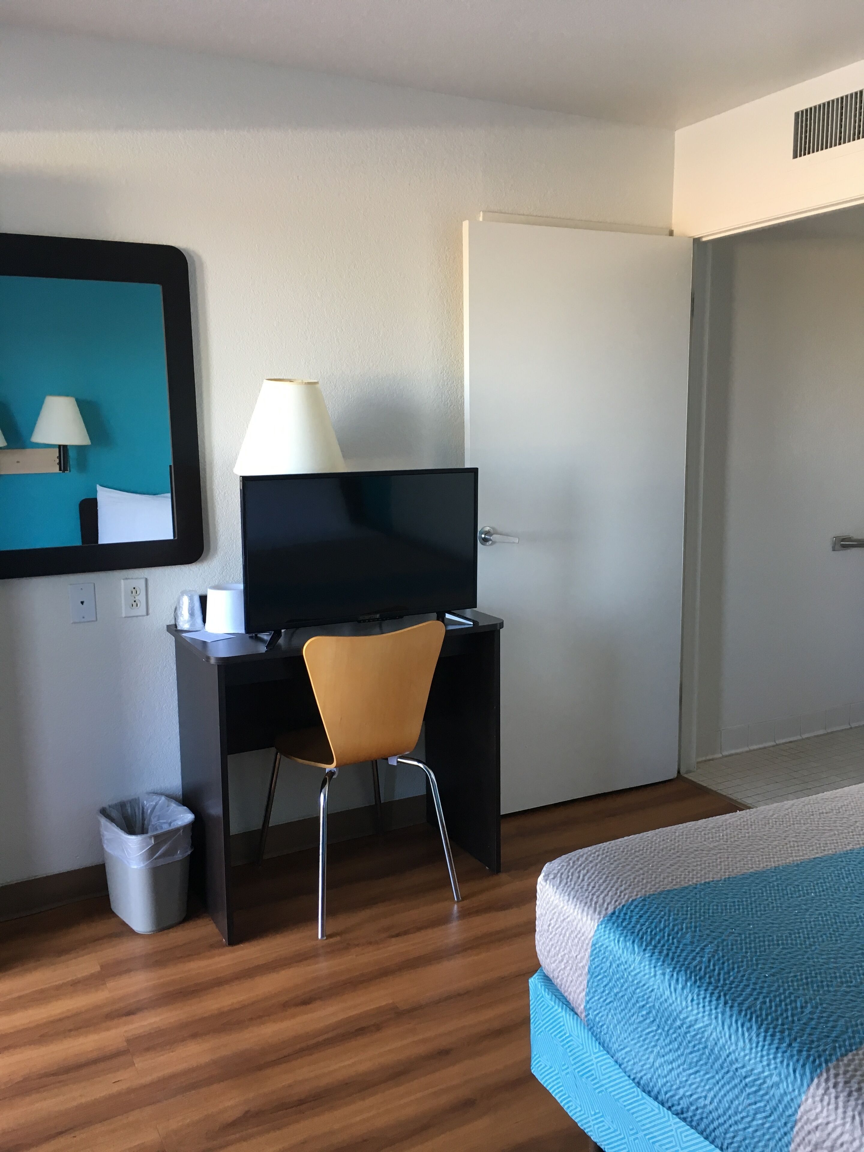 Habitación de hotel con cama, escritorio con computadora y lámpara, una silla, un cesto de basura y una puerta.