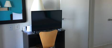 Habitación de hotel con cama, escritorio con computadora y lámpara, una silla, un cesto de basura y una puerta.