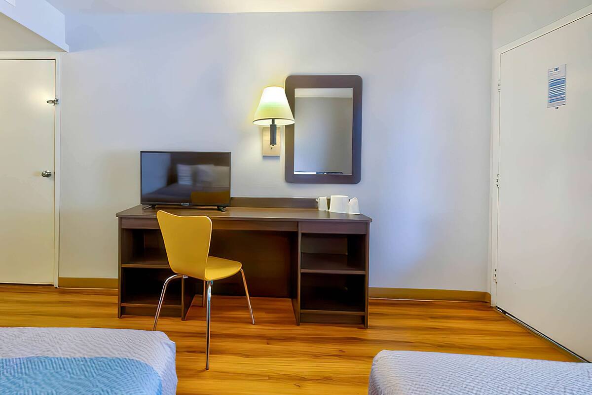 A hotel room with a wooden desk, a chair, a bed, a television, and a mirror.