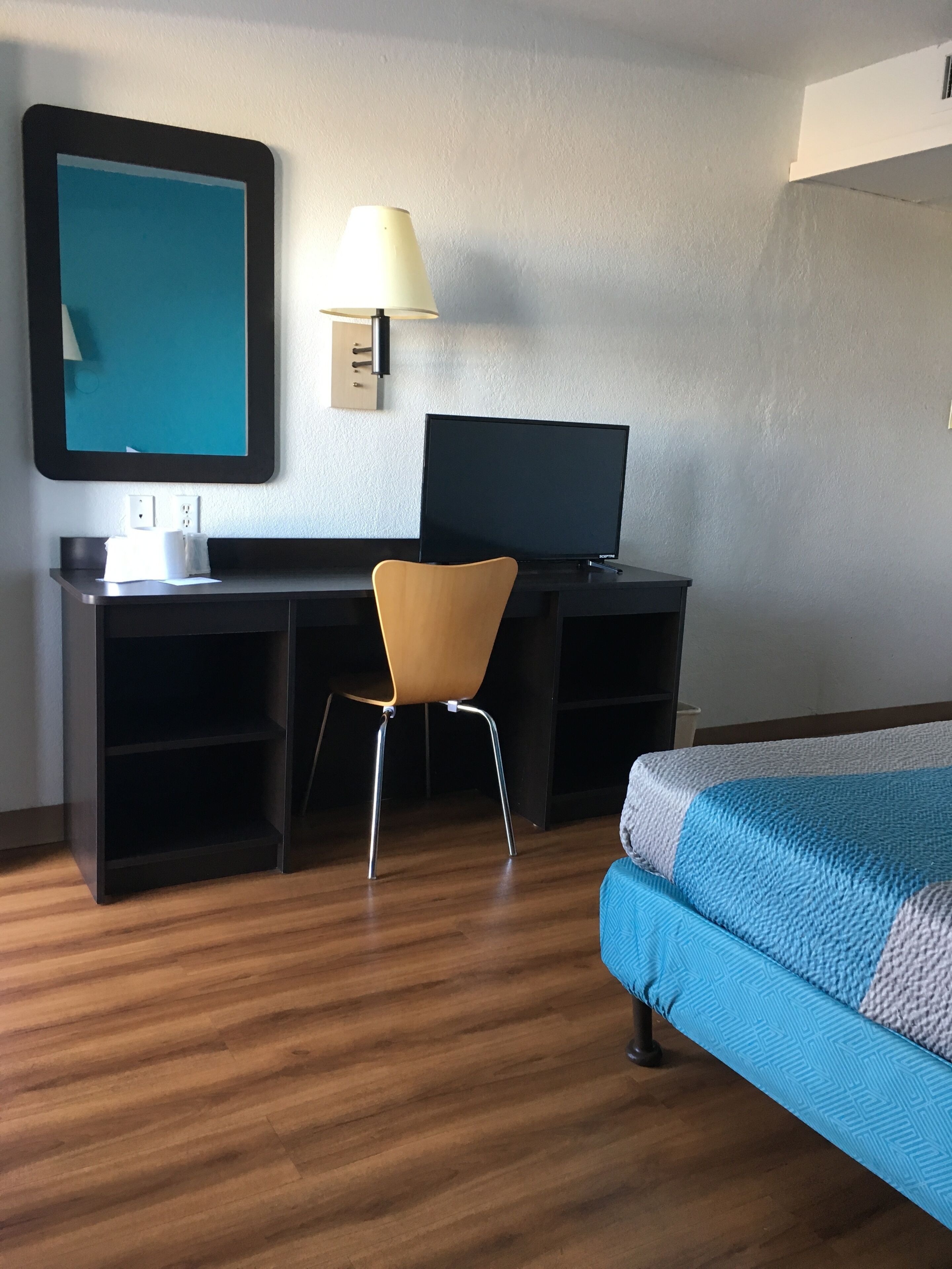A hotel room with a bed, a desk with a computer, a chair, a lamp, and a large mirror on the wall.