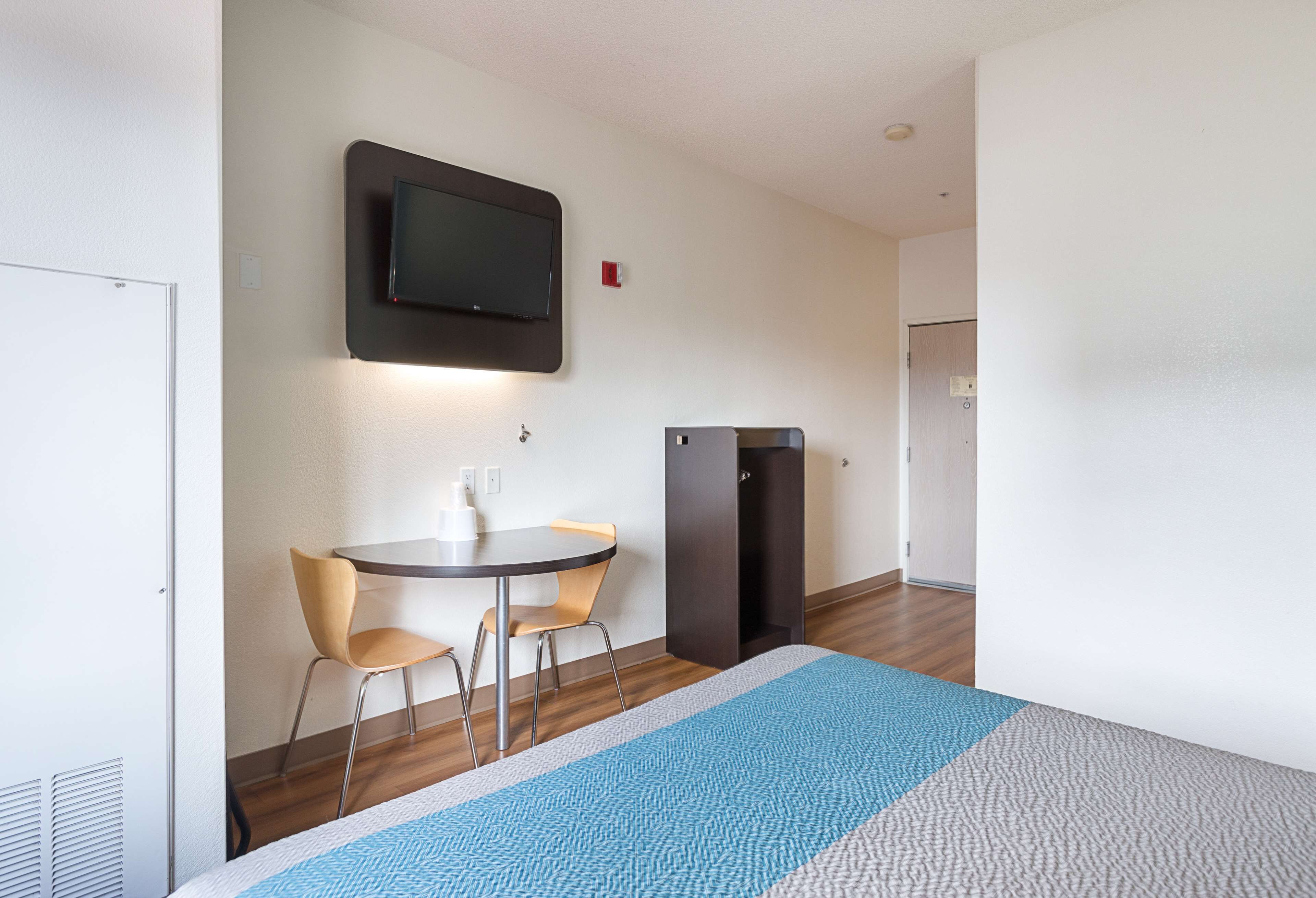 A hotel room with a bed, a TV mounted on the wall, a small table with two chairs, and a wardrobe.