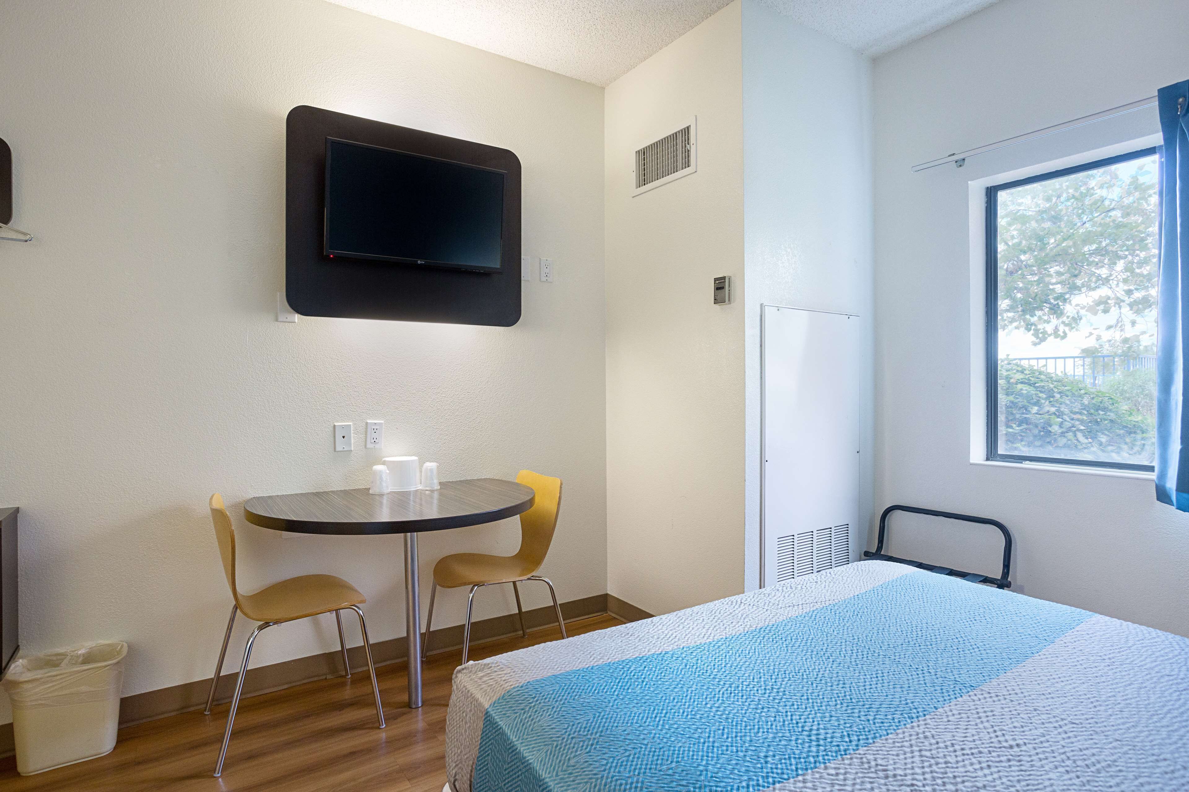 A hotel room with a bed, a TV mounted on the wall, a small table with two chairs, and a window with curtains.