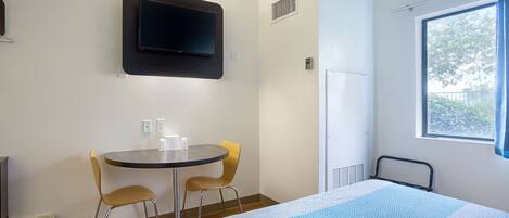 A hotel room with a bed, a TV mounted on the wall, a small table with two chairs, and a window with curtains.