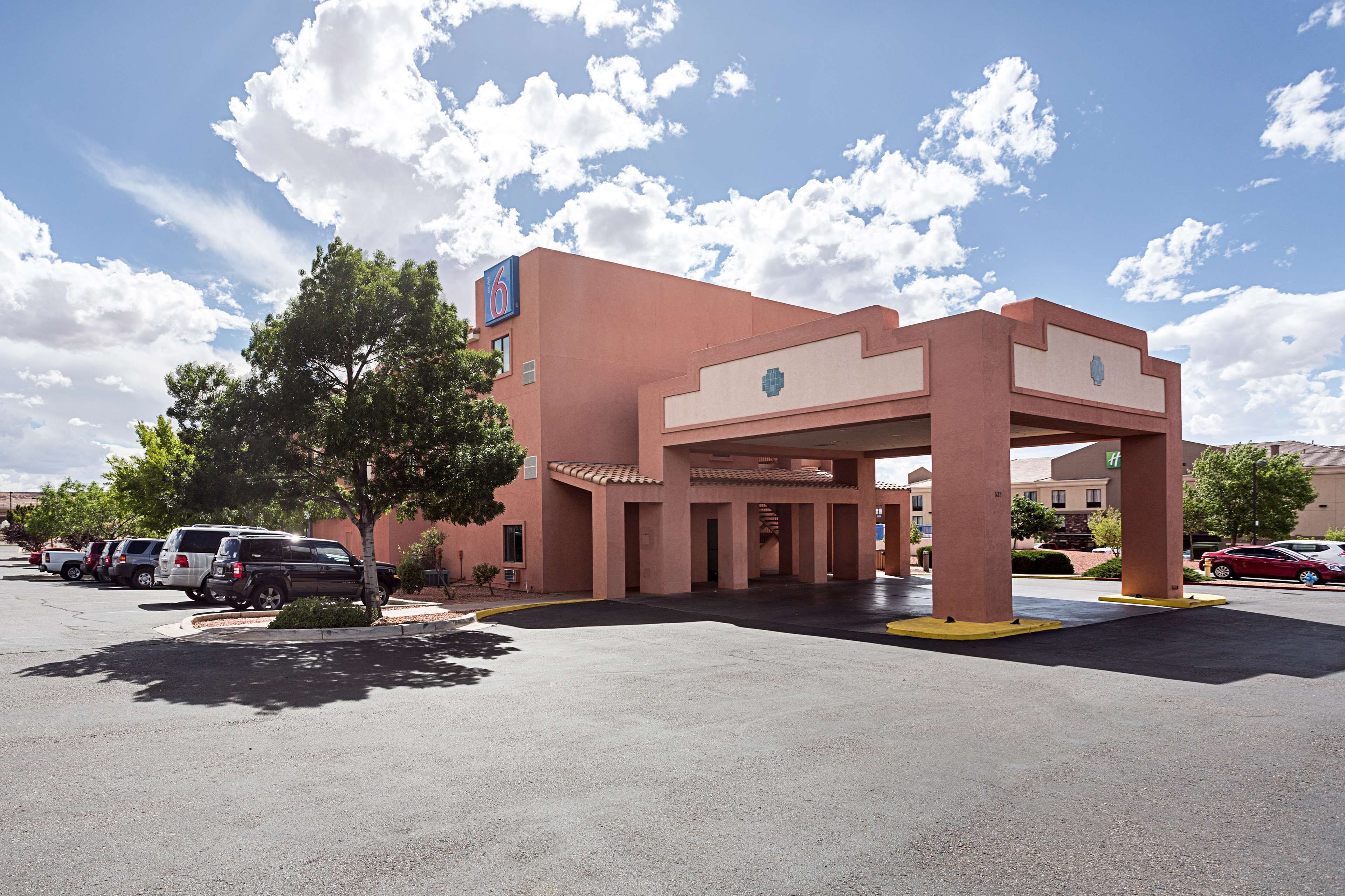 Un hotel con fachada roja, un acceso cubierto y un estacionamiento con varios coches.