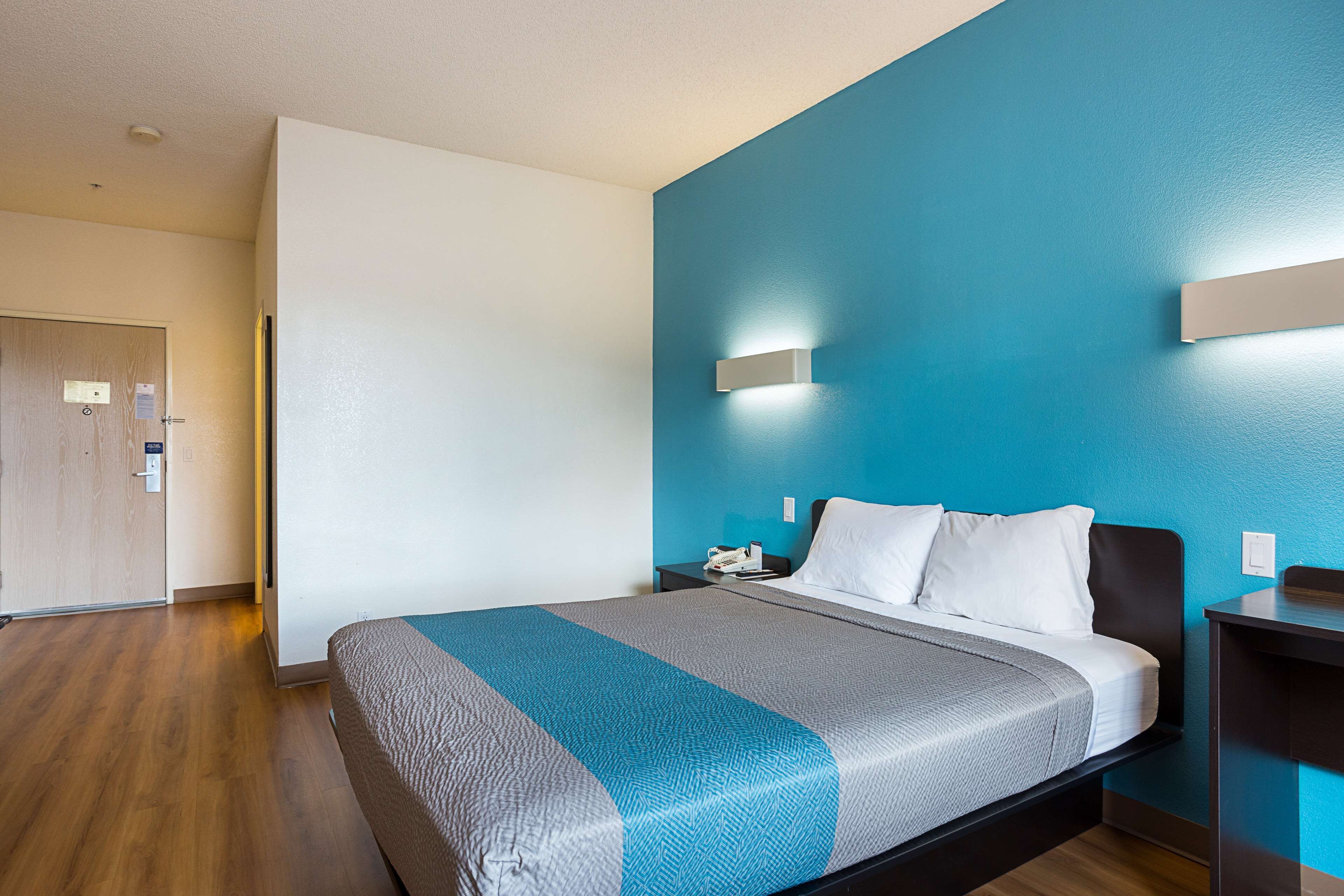 A hotel room with a bed, a nightstand, a wall-mounted light, and a door.