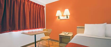 A hotel room with a wooden floor, a bed with white bedding, a small table with a chair, a nightstand with a lamp, and a window with red curtains.