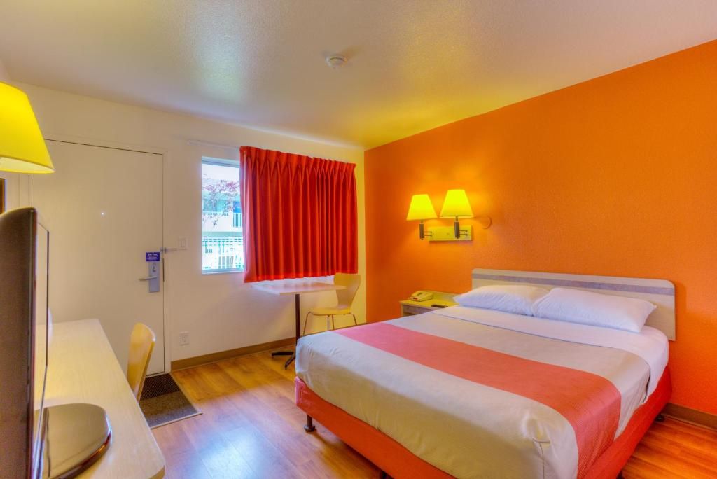 Une chambre d’hôtel avec un lit, un bureau, une chaise, une fenêtre avec des rideaux rouges et deux lampes de chevet.