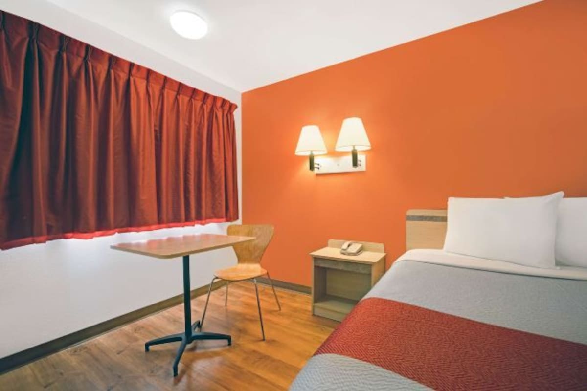 Habitación de hotel con cama, una mesita, dos sillas, una mesita de noche y una ventana con cortinas rojas.