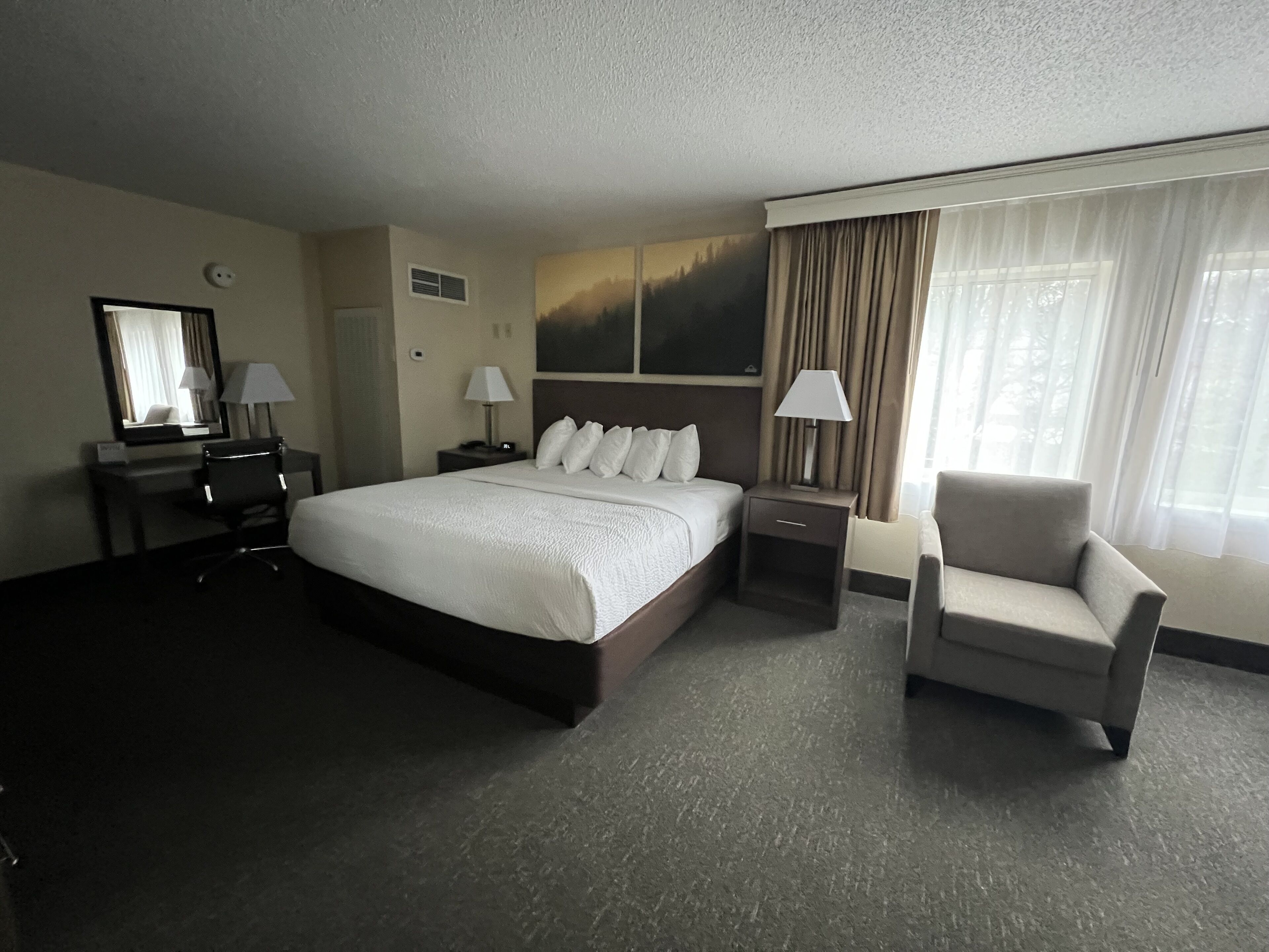 A hotel room with a large bed, a desk with a chair, a small table with a lamp, and a window with curtains.