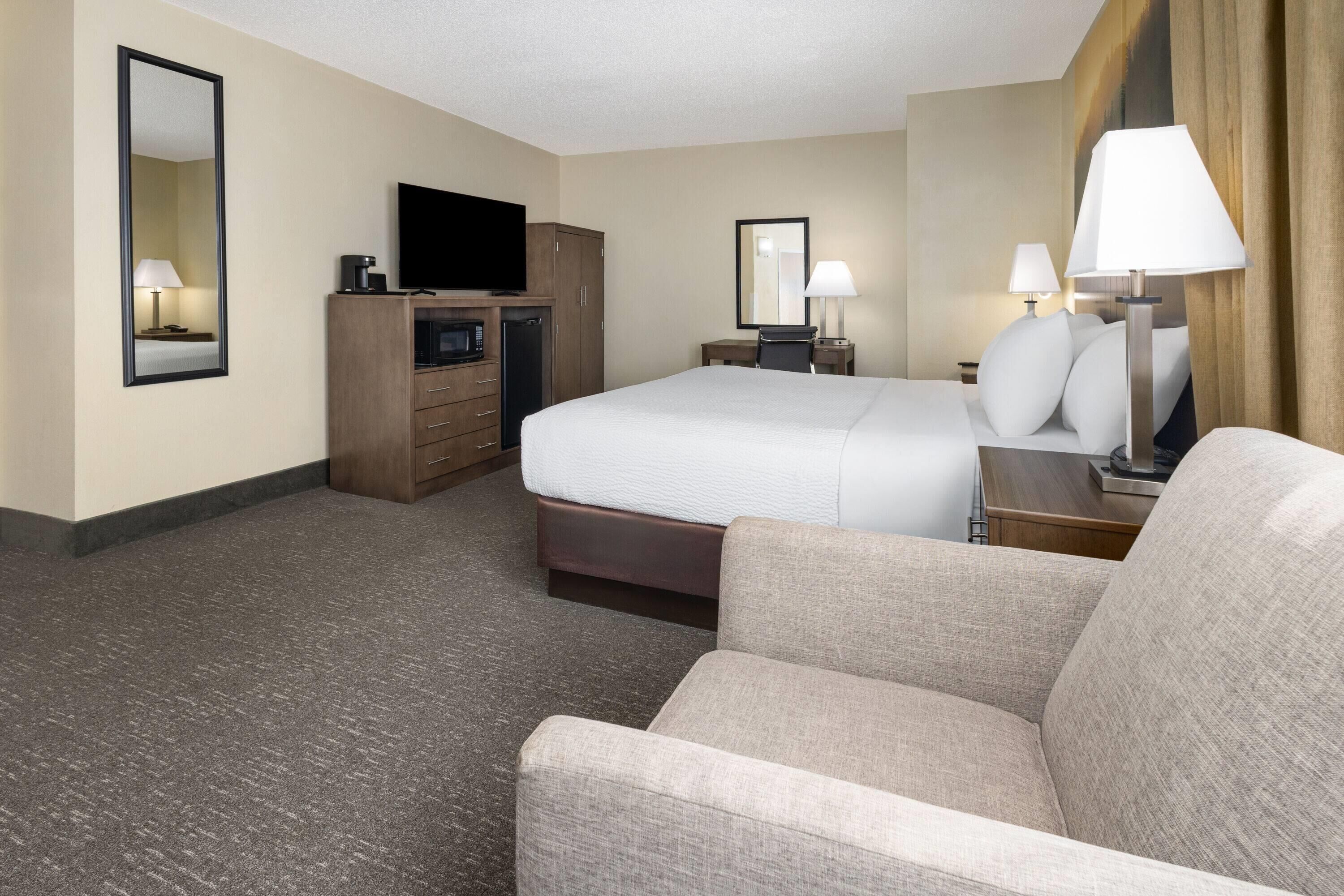 A hotel room with a bed, a TV, a desk with a lamp, a dresser, and a chair.