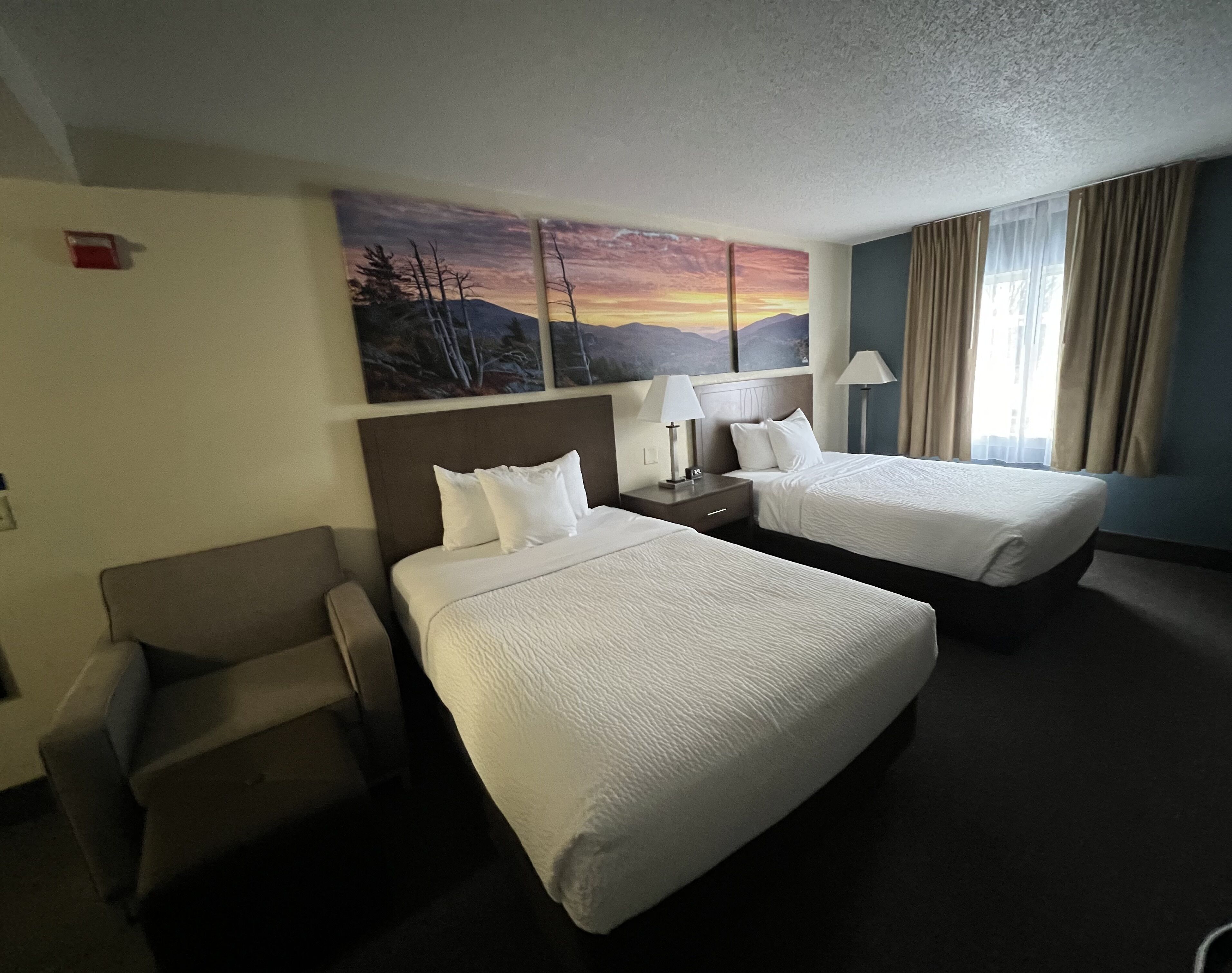 Habitación de hotel con dos camas, una silla, una mesita de noche, una lámpara y un cuadro de un atardecer.