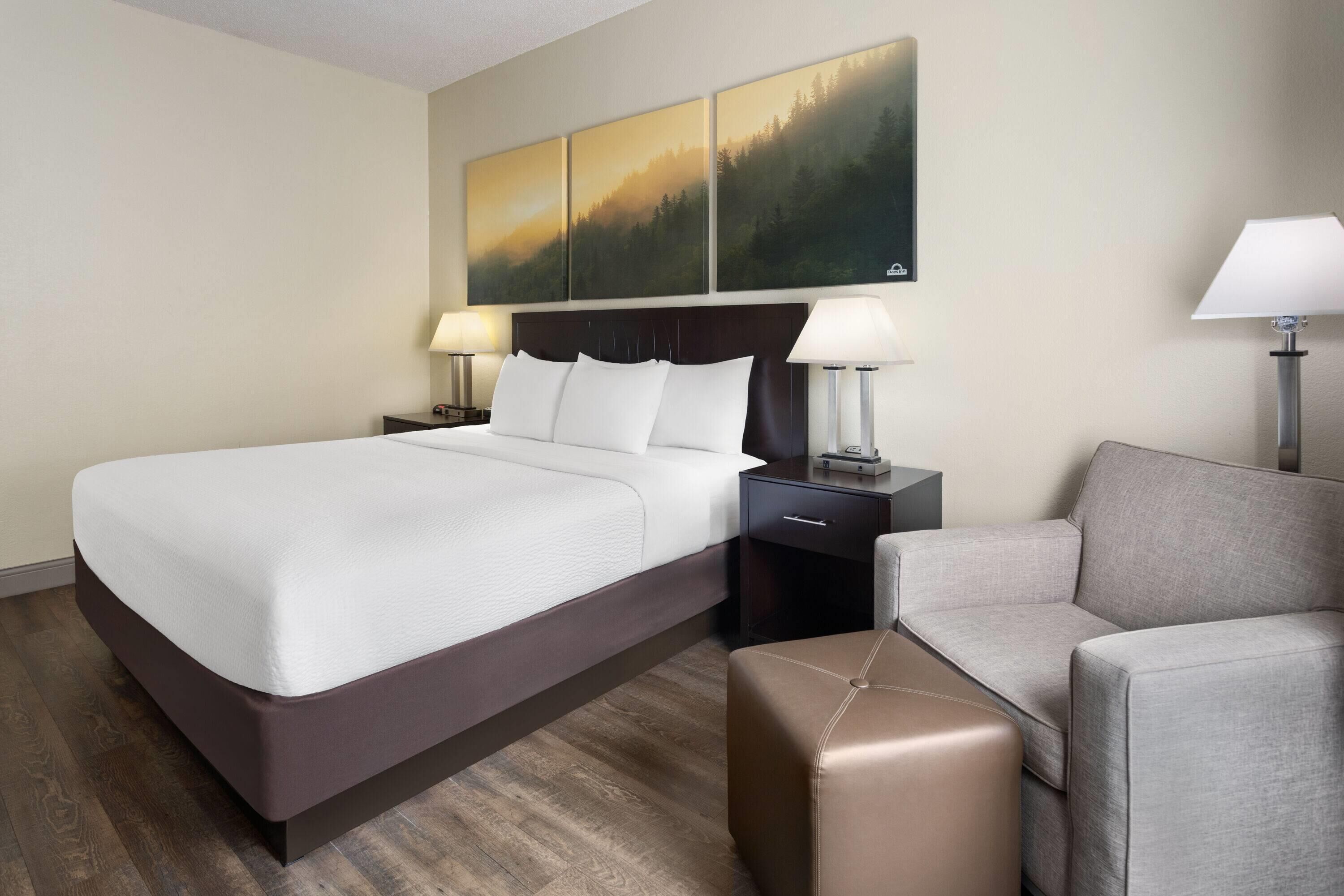 Une chambre d’hôtel avec un grand lit, deux tables de chevet avec des lampes, un canapé gris et un pouf marron.