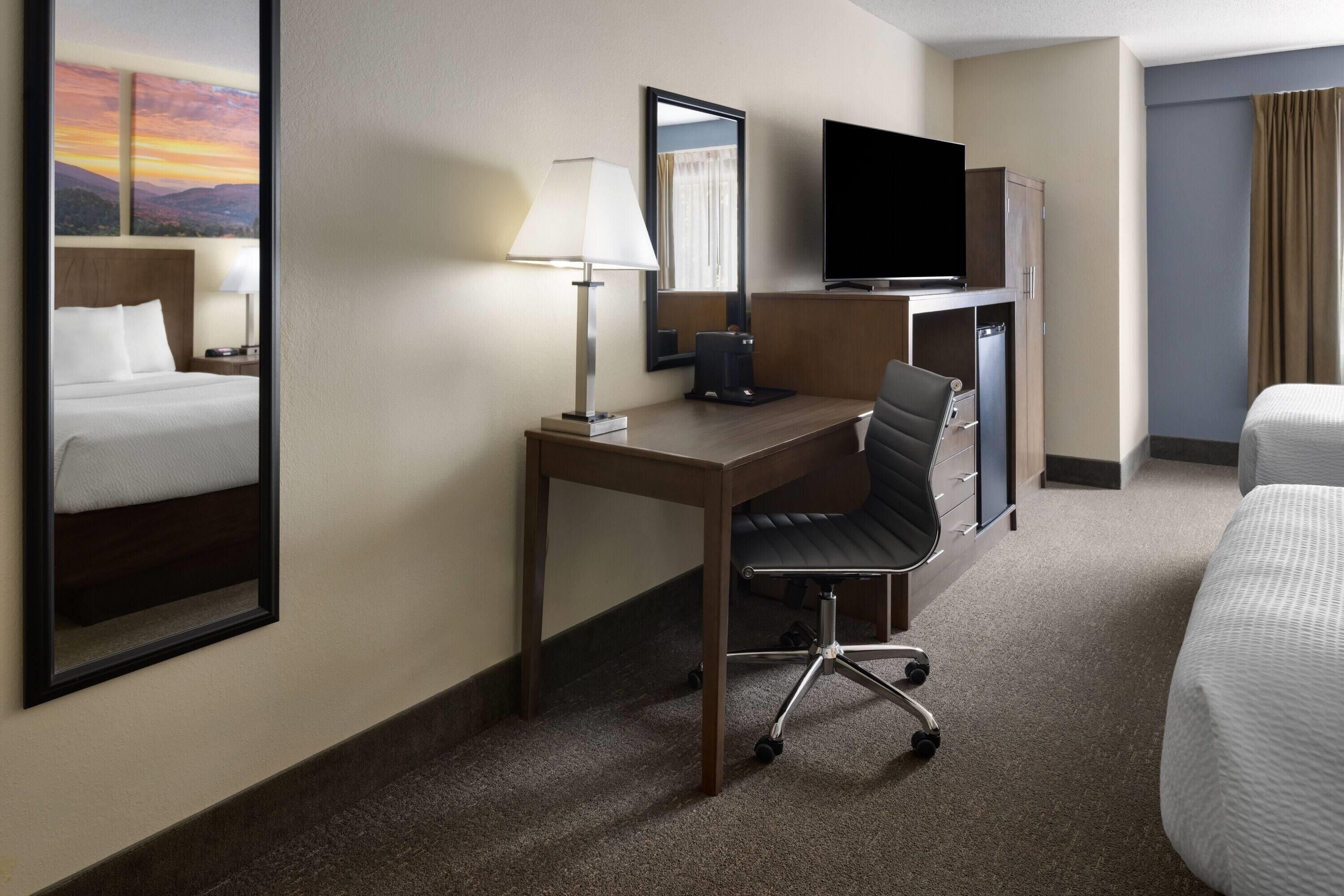 Una habitación de hotel con cama, escritorio, silla, televisor y espejo.
