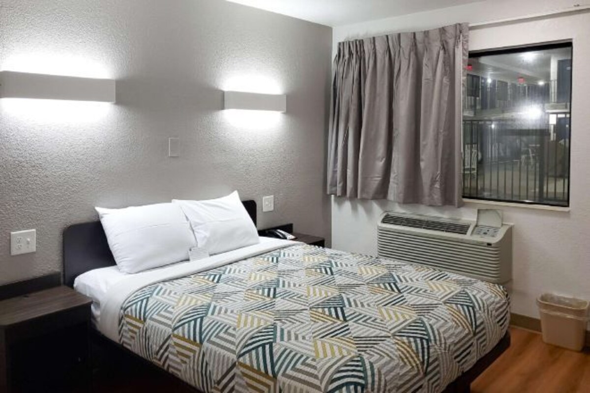 A hotel room with a bed, a window with curtains, an air conditioning unit, and a wall-mounted light fixture.