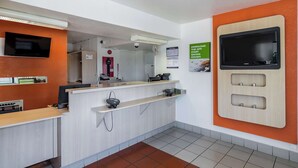 Un’area reception con un banco, un televisore a parete, una targa informativa e una presa elettrica fissata alla parete.