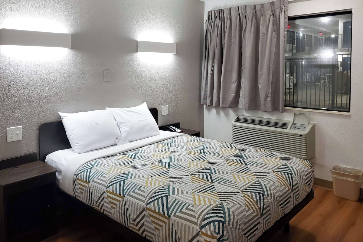 A hotel room with a bed, an air conditioning unit, a window with curtains, and a wall-mounted light fixture.