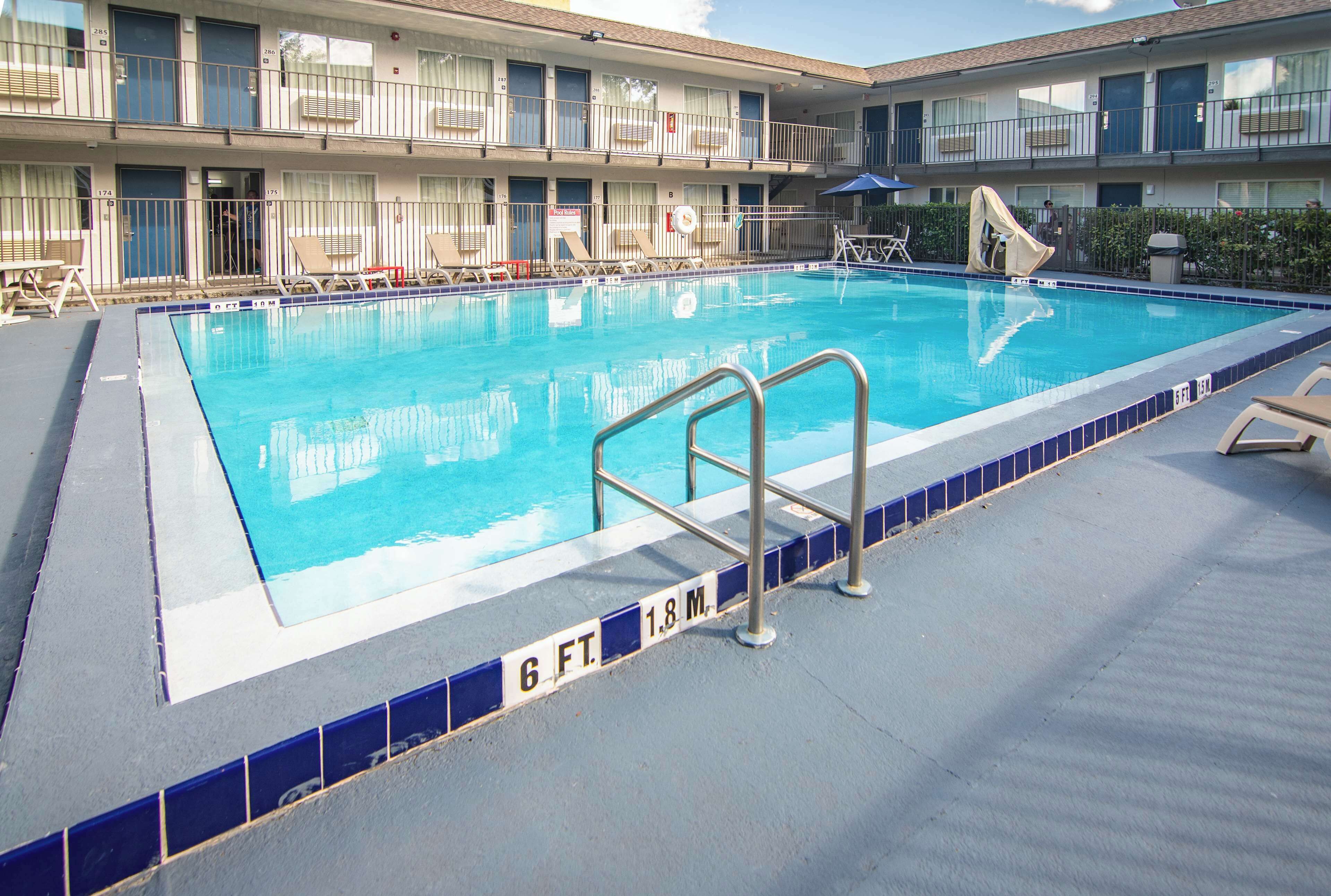 A swimming pool with a blue tiled border, lounge chairs, and a multi-story building in the background.