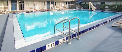 A swimming pool with a blue tiled border, lounge chairs, and a multi-story building in the background.