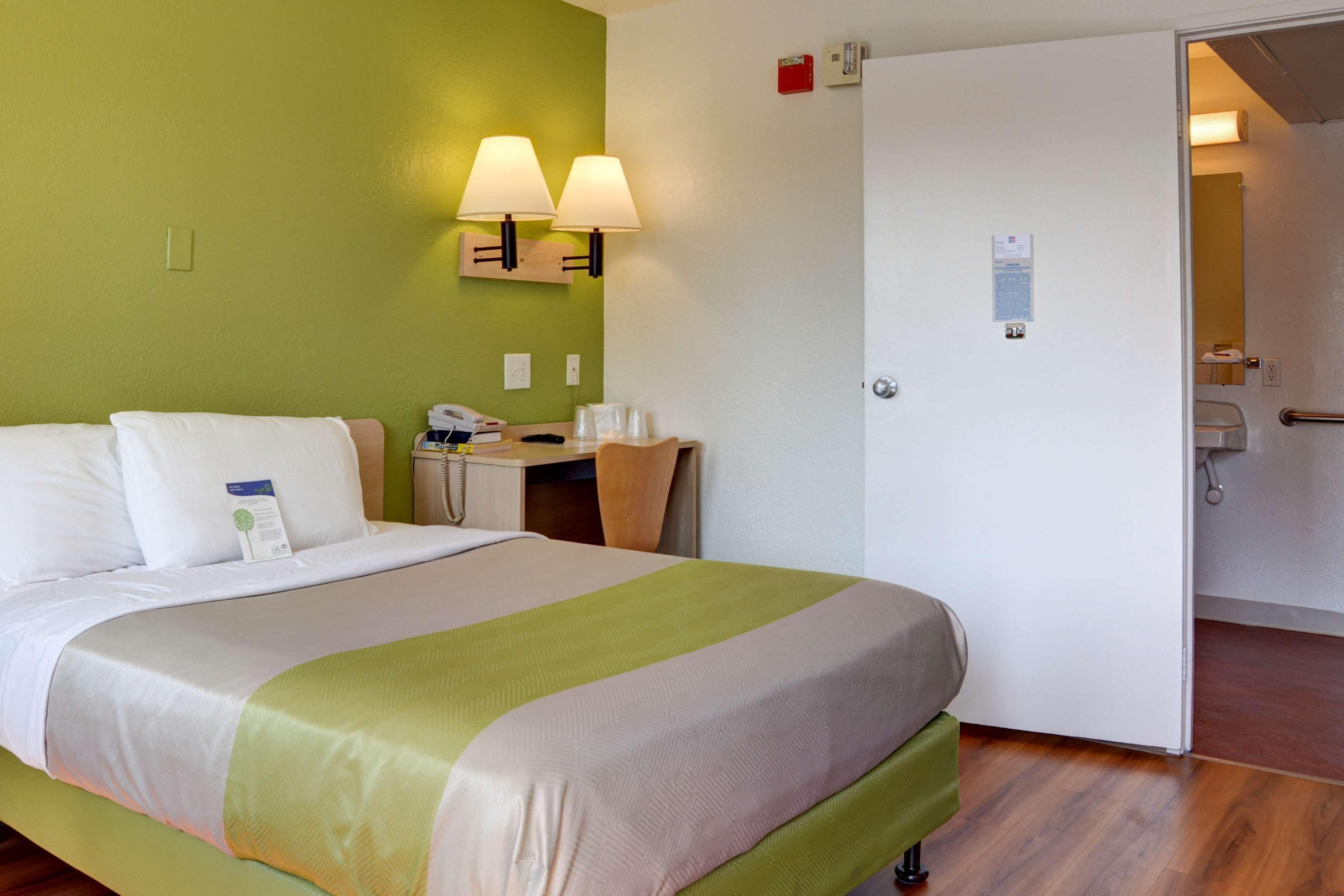 Habitación de hotel con cama, mesita de noche, lámpara y una puerta que da al baño.