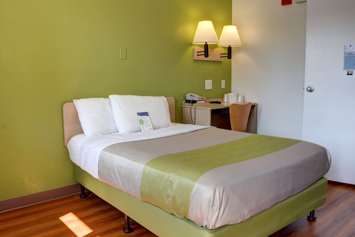Quarto de hotel com cama, mesa de cabeceira, abajur e parede verde.